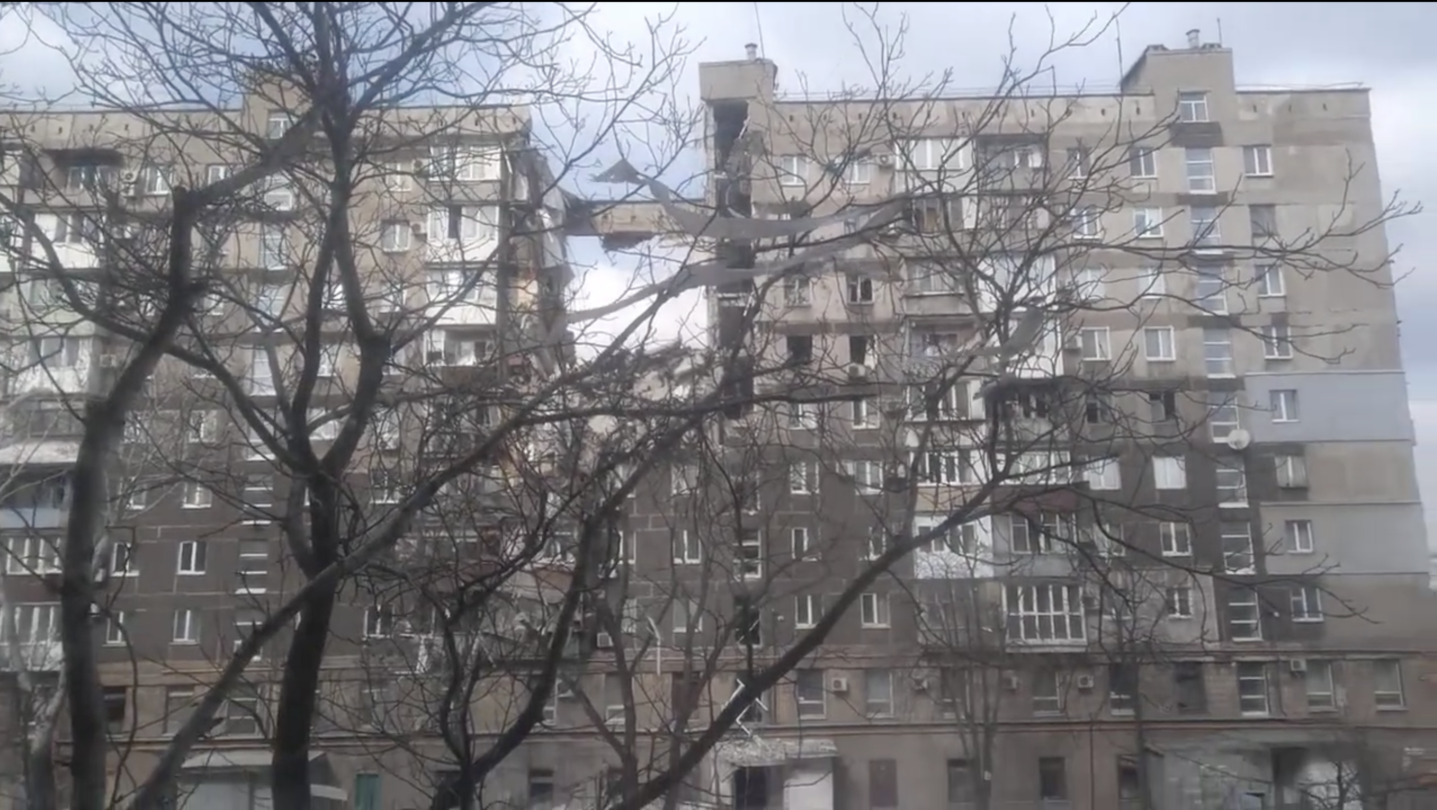 Street-level photograph, March 2022: the same nine-story building seen through bare winter trees, one entrance section collapsed clean through, exposing sky between the two remaining wings.